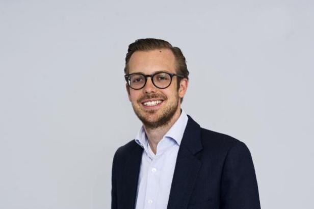 Ein lächelnder Mann mit Brille und Anzug vor einem hellen Hintergrund.