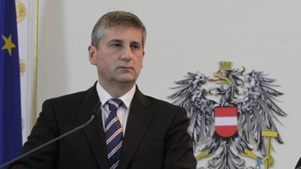 Ein Mann in Anzug und Krawatte steht vor der österreichischen Flagge und dem Bundeswappen.