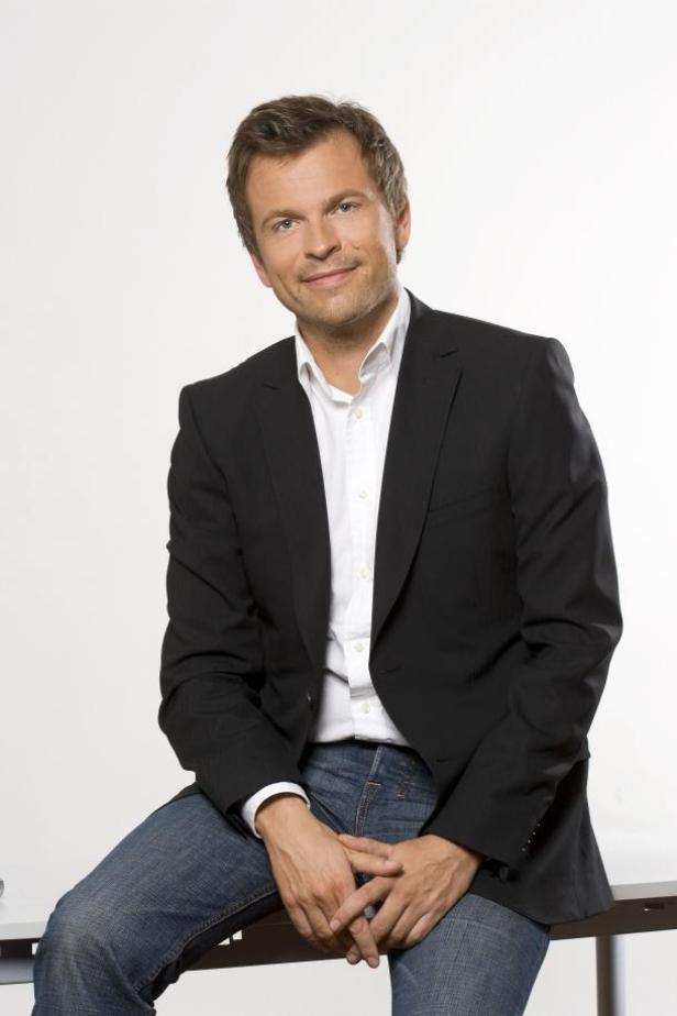 Ein Mann mit dunklem Sakko und Jeans sitzt lächelnd da.