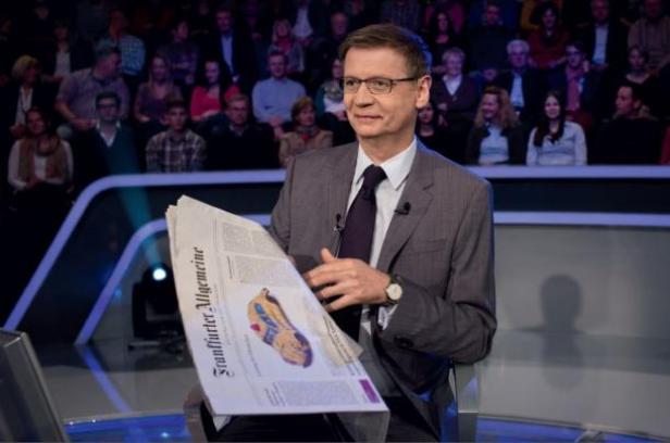 (c: f.a.z. - scholz & friends - matthias staetgens) Ein Mann im Anzug sitzt vor Publikum und liest die „Frankfurter Allgemeine Zeitung“.