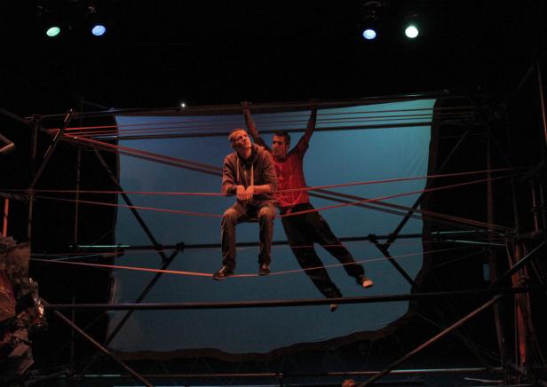 Zwei Männer auf einer Bühne, einer sitzend, der andere sich an einem Seil festhaltend.