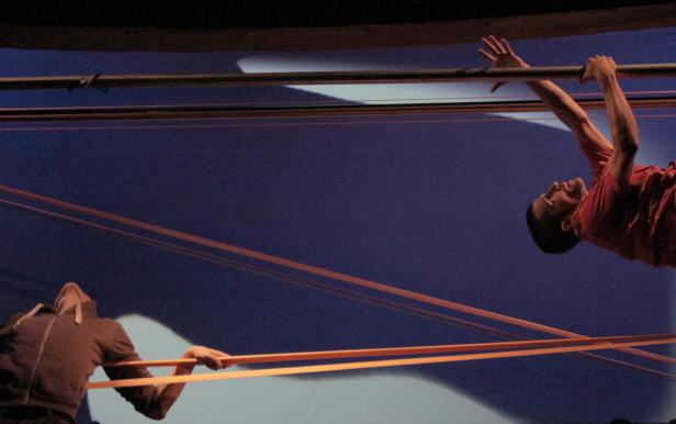 Zwei Männer performen an einem Gerüst mit gespannten Bändern vor einem blauen Hintergrund.