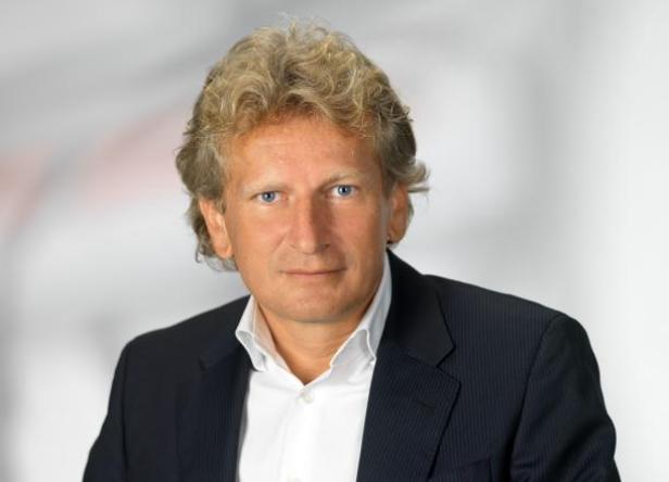 Porträt eines Mannes mit blonden Haaren und blauem Blazer vor einem grauen Hintergrund.