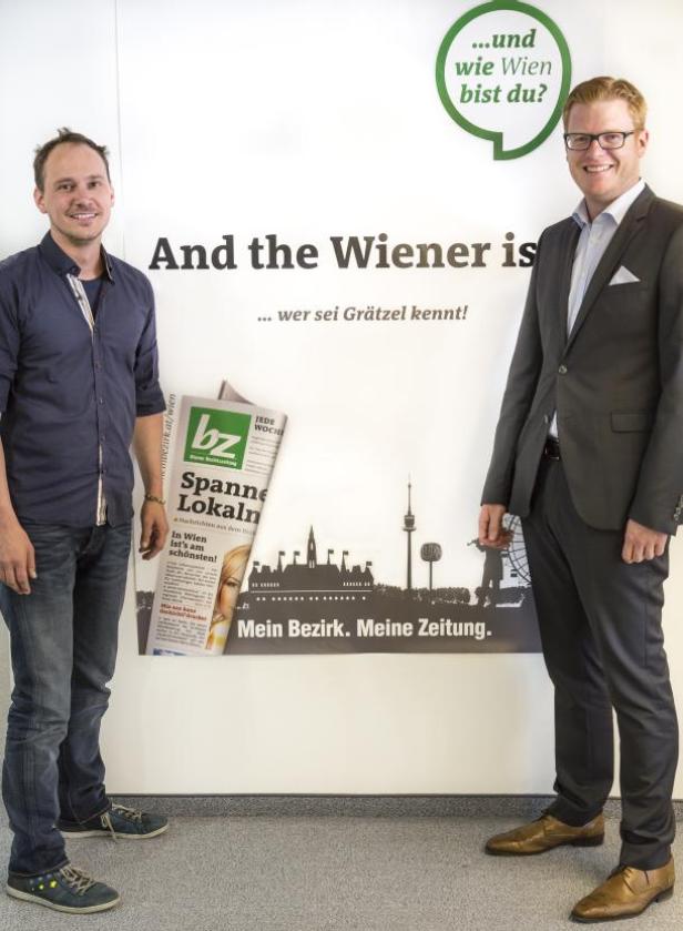 Zwei Männer posieren vor einem Werbeplakat der „Bezirkszeitung Wien“.