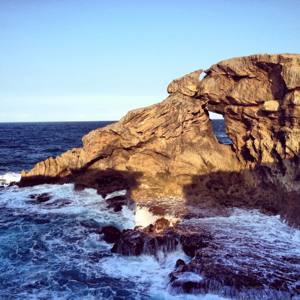 Eine Felsformation mit einem natürlichen Bogen steht an der Küste, umspült von Wellen.