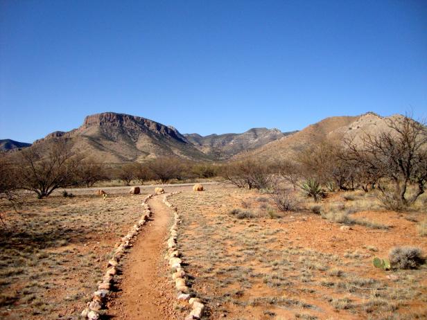 Ein sandiger Pfad führt durch eine trockene Landschaft mit Bergen im Hintergrund.