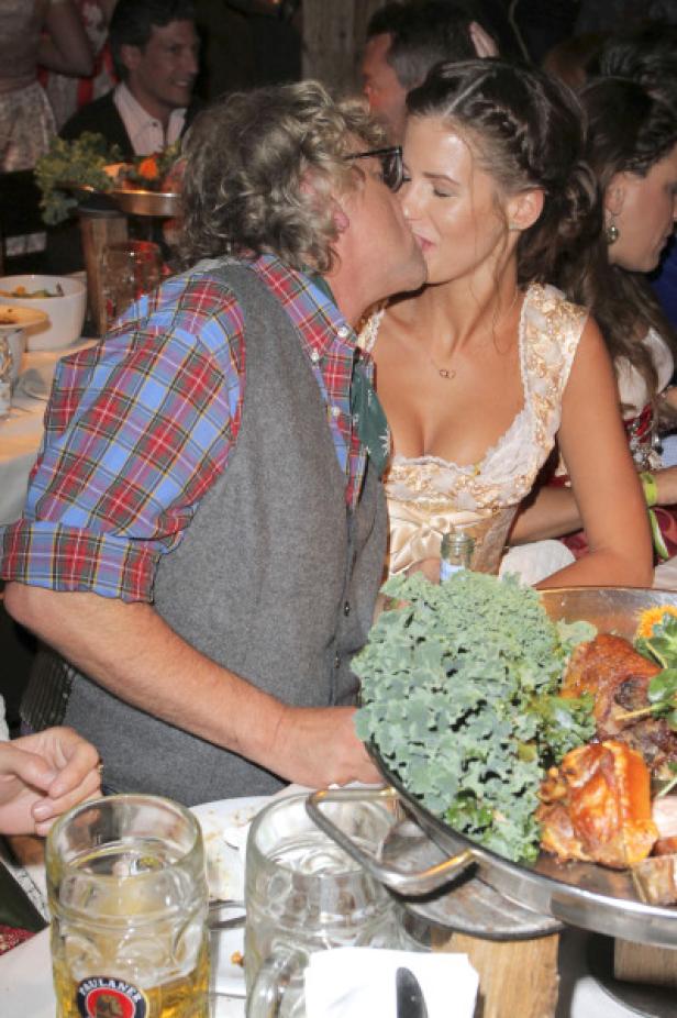 Ein Mann küsst eine Frau in Dirndl vor einem Tisch mit Speisen und Bier.