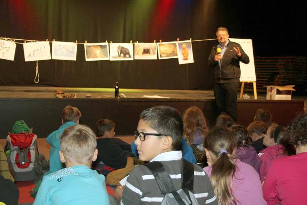 Ein Mann hält einen Vortrag vor einer Gruppe von Kindern auf einer Bühne.
