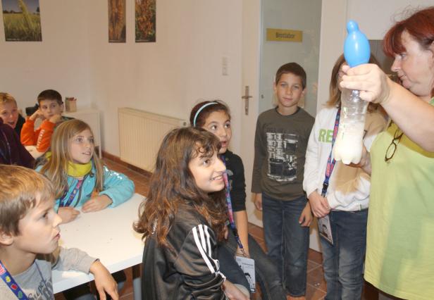Eine Frau führt Kindern in einem Raum ein Experiment mit einer Flasche und einem Ballon vor.