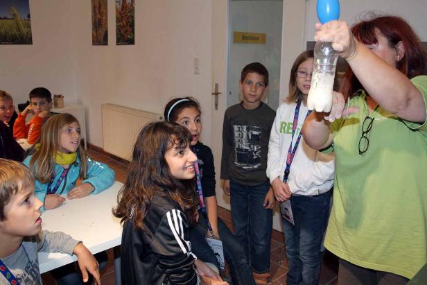 Eine Frau führt ein Experiment mit einer Flasche und einem Ballon vor einer Gruppe von Kindern durch.