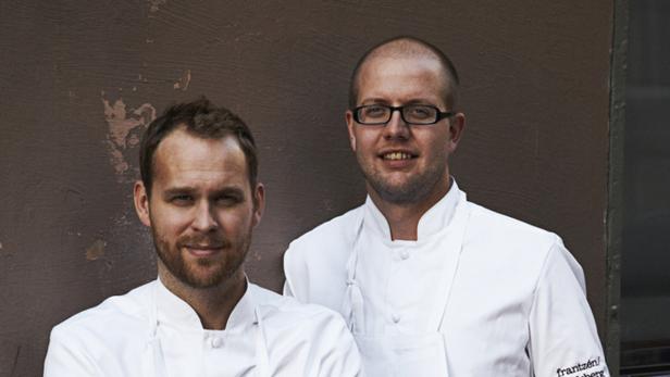 Zwei Köche in Kochjacken stehen vor einer braunen Wand.