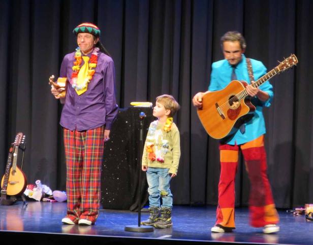 Zwei Clowns und ein Kind stehen auf einer Bühne, einer spielt Gitarre, der andere hält eine Ukulele.