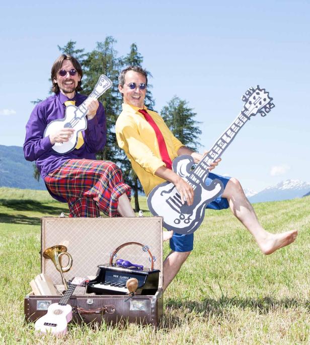 Zwei Männer posieren mit Pappgitarren und Musikinstrumenten vor einer Bergkulisse.