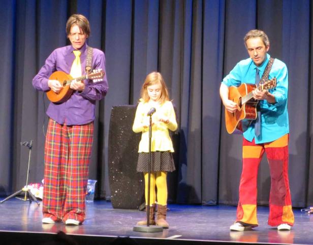 Zwei Männer in bunten Kostümen spielen Gitarre, während ein junges Mädchen am Mikrofon steht.