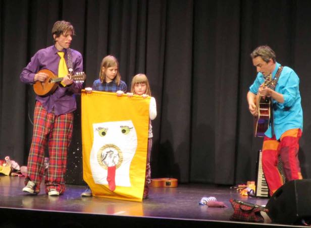 Zwei Musiker und zwei Kinder treten mit einem Banner auf einer Bühne auf.