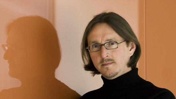 Ein Mann mit Brille und schwarzem Rollkragenpullover steht vor einer Wand.