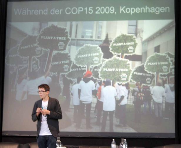 Ein Redner steht vor einer Projektion der COP15 in Kopenhagen, auf der Demonstranten mit „Plant a Tree“-Schildern zu sehen sind.
