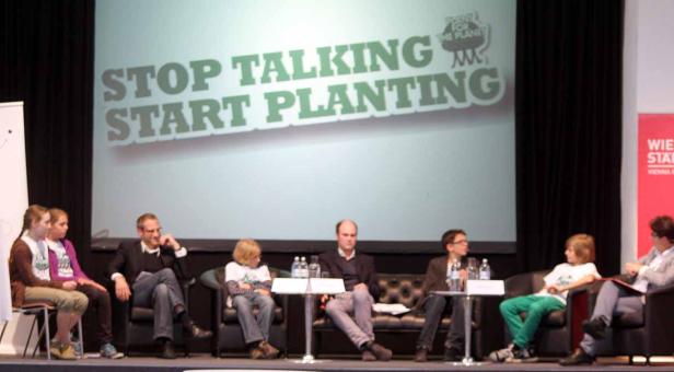 Eine Gruppe von Menschen sitzt auf einer Bühne vor einem Bildschirm mit der Aufschrift „Stop Talking Start Planting“.