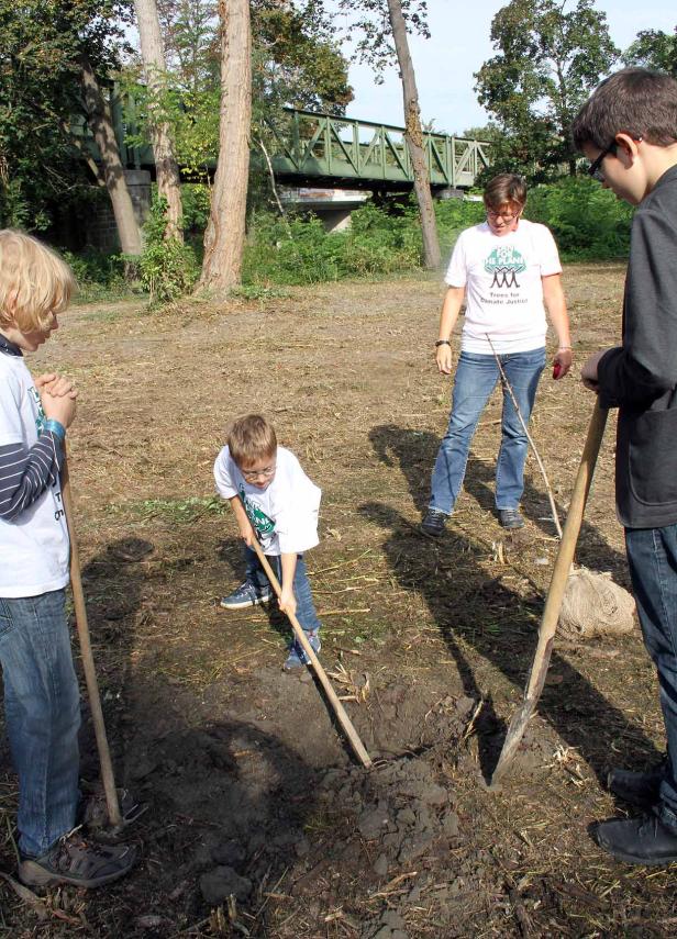 Eine Gruppe von Kindern und Erwachsenen pflanzt einen Baum mit Spaten in einem Park.