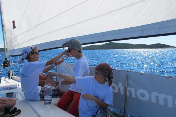 Drei Jugendliche mit Kopftüchern sitzen auf einem Segelboot auf dem glitzernden Meer.