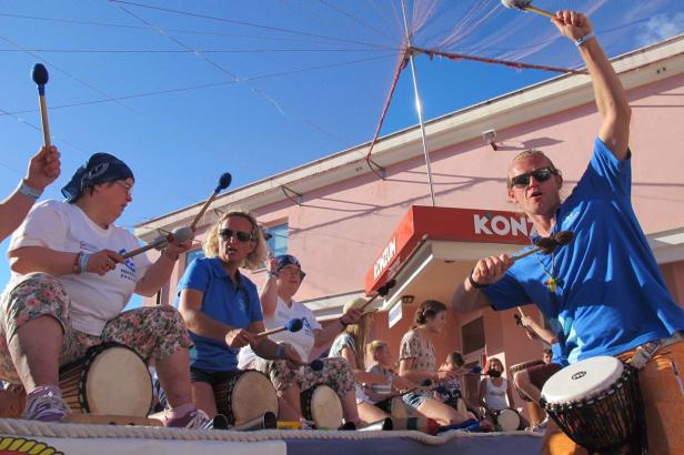 Eine Gruppe von Menschen spielt auf Djemben vor einem Geschäft mit der Aufschrift „Konzum“.