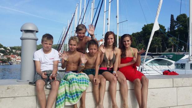 Eine Gruppe Jugendlicher sitzt an einem sonnigen Tag am Hafen und winkt.