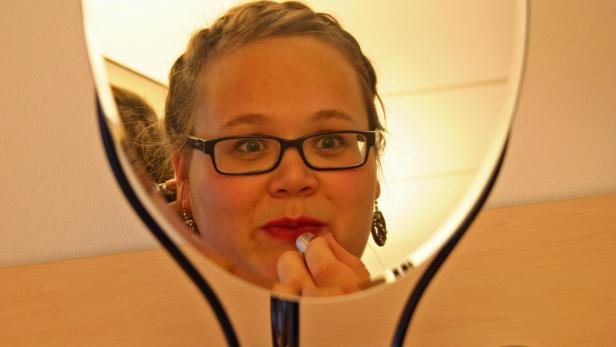 Eine junge Frau mit Brille trägt Lippenstift vor einem Spiegel auf.