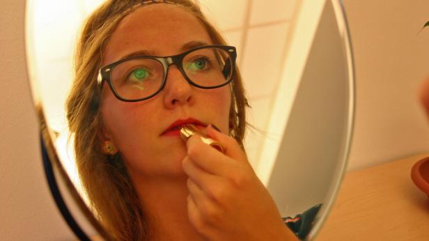 Eine junge Frau mit Brille trägt Lippenstift vor einem Spiegel auf.