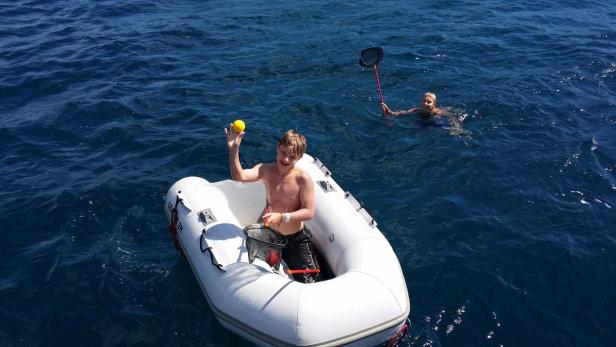 Ein Junge in einem Schlauchboot hält einen gelben Ball hoch, während ein anderer im Wasser mit einem Kescher steht.