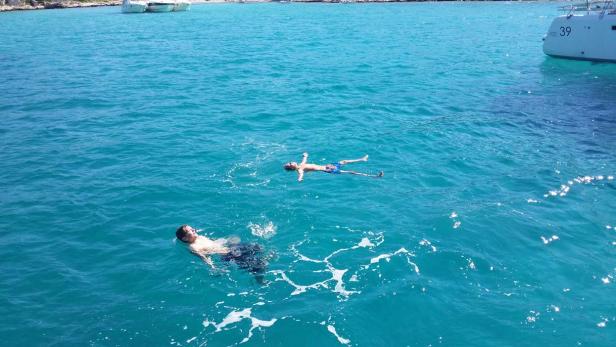 Zwei Personen schwimmen im türkisfarbenen Meer, im Hintergrund ein Segelboot.