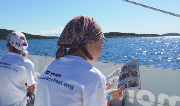 Eine Person mit Kopftuch liest ein Comicbuch auf einem Boot mit Blick auf das Meer.
