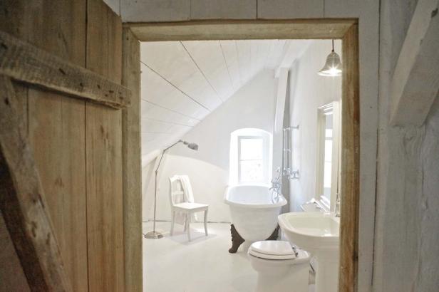 Blick durch eine Holztür in ein weißes Badezimmer mit Badewanne und Dachschräge.