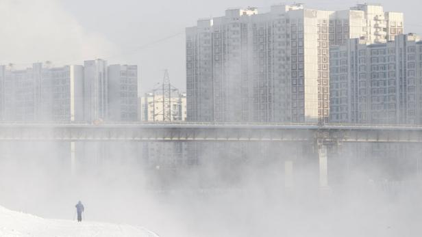 Eine Person geht im Winter an einem Fluss mit Hochhäusern im Hintergrund spazieren.