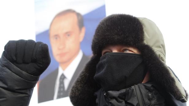 Eine vermummte Person mit geballter Faust vor einem Porträt von Wladimir Putin.