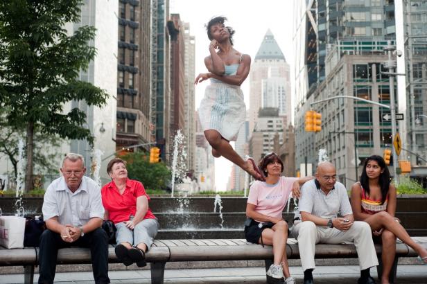 Eine Frau springt vor einem Brunnen in der Stadt, während andere auf einer Bank sitzen.