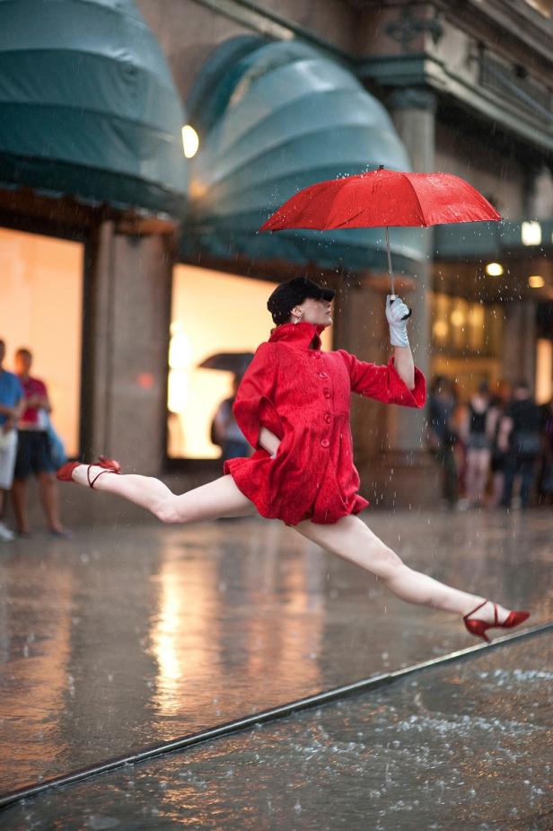Eine Frau in rotem Mantel und Schuhen springt mit einem roten Regenschirm im Regen.