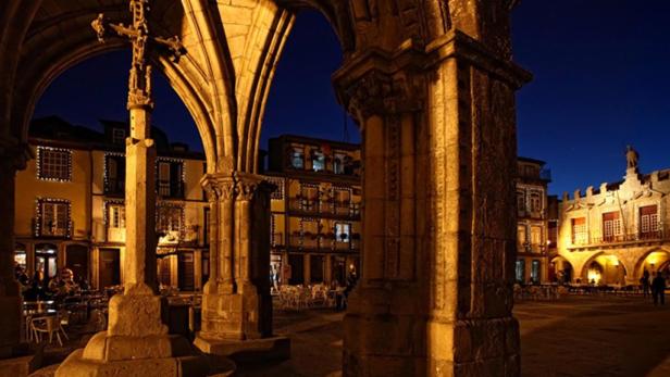 Ein beleuchteter Platz mit historischen Gebäuden und einem steinernen Kreuz bei Nacht.