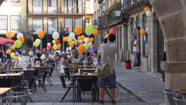 Ein belebter Platz mit vielen Menschen, Tischen, Stühlen und bunten Ballons.