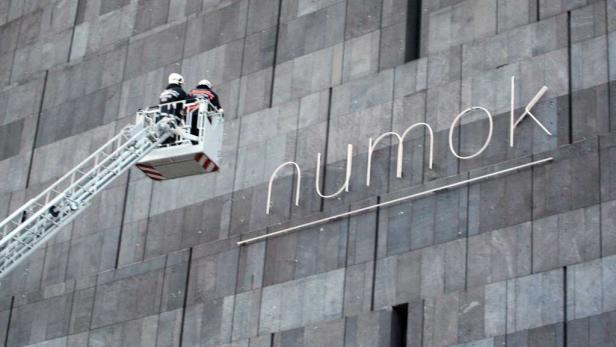 Zwei Arbeiter auf einer Hebebühne montieren den Schriftzug „numok“ an einer grauen Fassade.