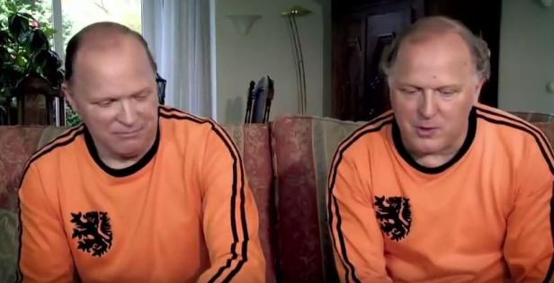 Zwei Männer, vermutlich Zwillinge, sitzen nebeneinander und tragen orangefarbene Trikots mit einem Löwenemblem.