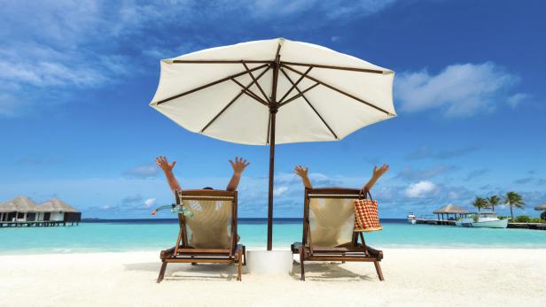 Zwei Personen entspannen unter einem Sonnenschirm am Strand mit türkisfarbenem Wasser.
