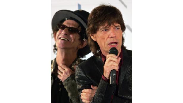 Mick Jagger spricht in ein Mikrofon, während Keith Richards im Hintergrund lächelt.