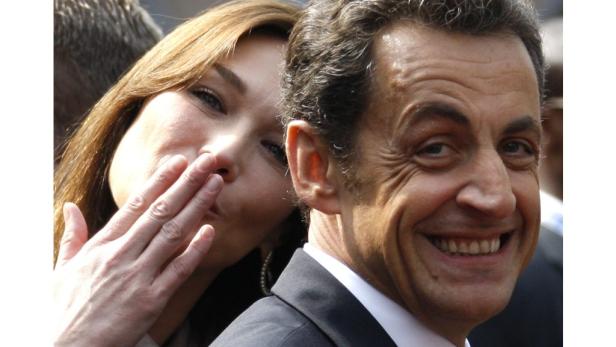 Nicolas Sarkozy lächelt, während Carla Bruni ihm einen Kuss zuhaucht.