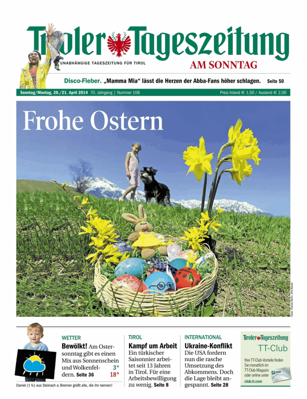 Ein Osterkorb mit bunten Eiern, einem Stoffhasen und Narzissen im Gras vor einer Bergkulisse.