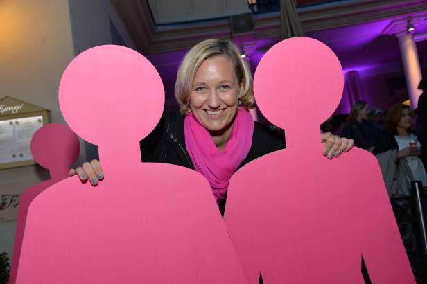 Eine lächelnde Frau posiert hinter zwei rosafarbenen Pappfiguren.