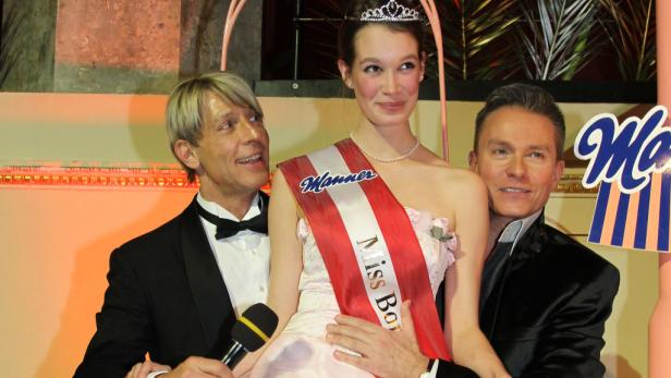 Eine junge Frau mit Krone und Schärpe „Miss Bonbon“ wird von zwei Männern im Smoking flankiert.