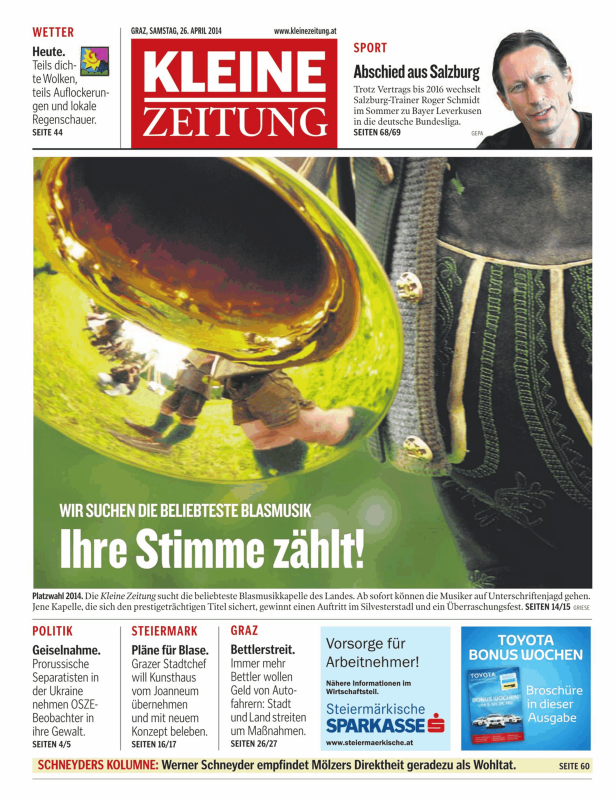 Eine Blaskapelle in Tracht spielt auf einer Wiese vor dem Hintergrund der „Kleine Zeitung“.