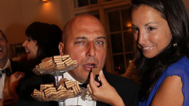 Eine Frau füttert einen Mann mit Pralinen, während er ein Auge zukneift.