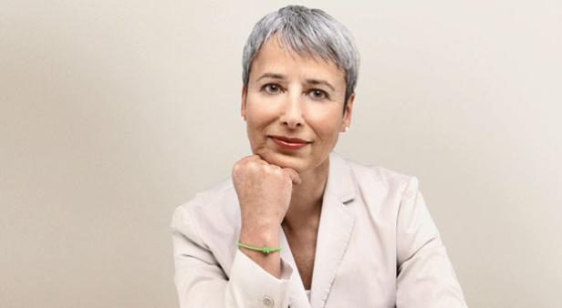 Eine Frau mit grauen Haaren in einem hellen Blazer posiert mit ihrer Hand am Kinn.