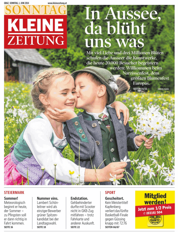 Ein Junge im Trachtenanzug gibt einem Mädchen einen Kuss auf die Wange, beide halten Narzissen.
